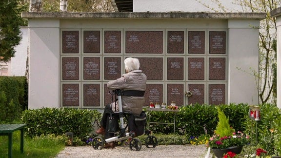 Eine weißhaarige Frau sitzt auf einem Rollator auf einem Friedhof vor einer Wand mit Inschriften.