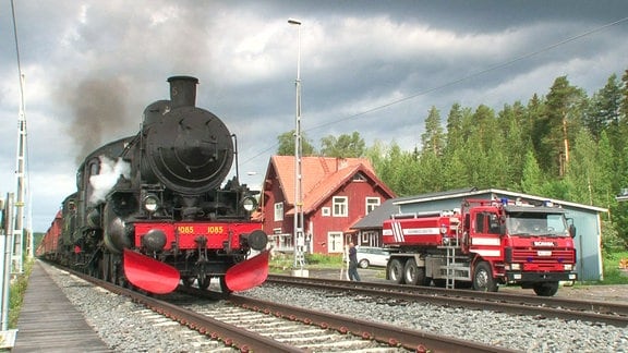 Eine alte Dampflok und ein Löschfahrzeug stehen vor einem rotgeestrichenen Haus an einem Wald.