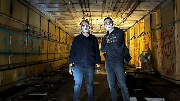 Zwei Personen stehen in einem langen Gang eines Bunkers mit Taschenlampen.