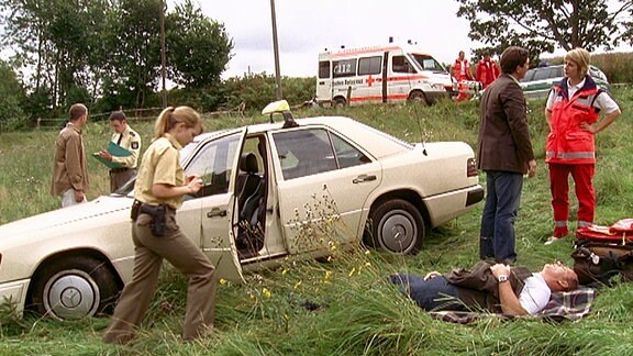 Sanitäter , Polizei und ein Mann vor einem Auto im Gras liegend