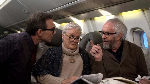 Eine Frau und ein Mann sitzen in einem Flugzeug. Im Gang steht ein Mann, der sich nah zu ihnen herüberbeugt und etwas zu ihnen sagt. Die beiden Sitzenden schauen ihn genervt bis wütend an. 