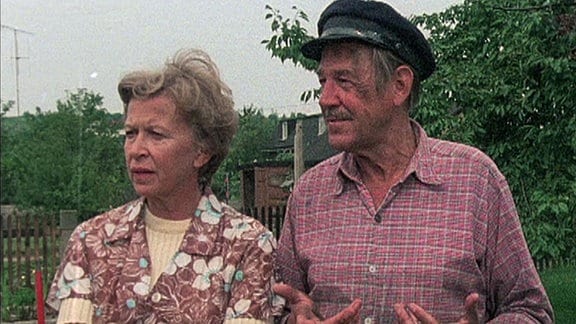 Eine Frau in Schürze und ein Mann mit Mütze stehen in einem Kleingarten.