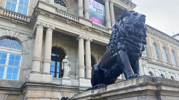 Eine Löwenskulptur sitzt vor einem klassizistischen Gebäude.