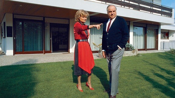 Hannelore und Helmut Kohl stehen vor einem Wohnhaus im Garten. Sie deutet auf einen Punkt außerhalb des Bildausschnitts, er blickt in die entsprechende Richtung.