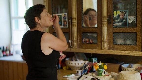 Eine Frau schminkt sich vor dem Spiegel eines alten Küchenschrankes.