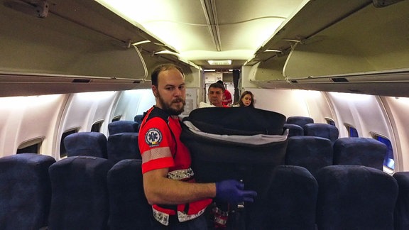 Ein Sanitäter trägt einen Korb mit einem Neugeborenen durch ein Flugzeug.