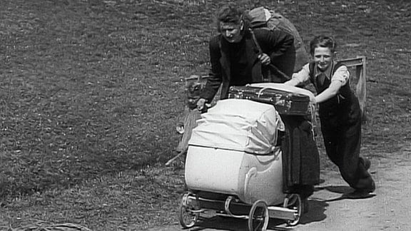 Ein Schwarzweißfoto, auf dem eine bepackte Mutter und ein Junge, der einen Kinderwageen schiebt, zu sehen sind.