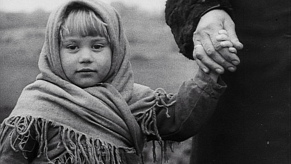 Ein Schwarzweißfoto, auf dem ein Mädchen mit Kopftuch, an einer Hand geht. 