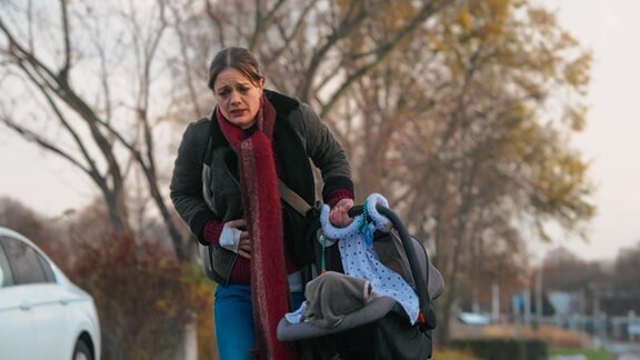 Eine junge Frau mit Babyschale in der Hand, hält sich mit schmerzverzerrtem Gesicht den Bauch.