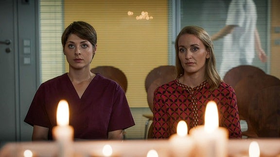 Zwei Frauen sitzen nebeneinander in einem Andachtsraum eines Krankenhauses vor brennenden Kerzen. 