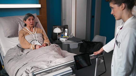 Eine ältere Frau in einem Krankenhausbett spicht mit einer jungen Ärztin.