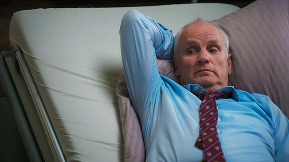 Ein Mann mit Hemd und Krawatte liegt auf einem Krankenbett.