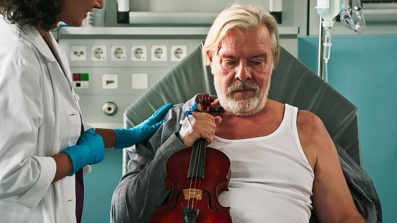 Ein Mann mit einer Geige in der Hand wird auf einer Behandlungsliege untersucht. 