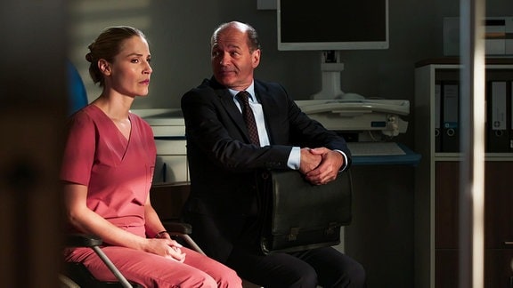 Eine Ärztin und ein Mann im Anzug sitzen nebeneinander in einem abgedunkelten Raum.
