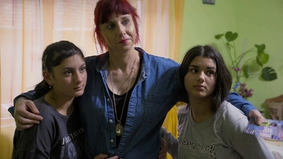 Eine junge Frau mit zwei Teenagern in den Armen steht in einem Zimmer.