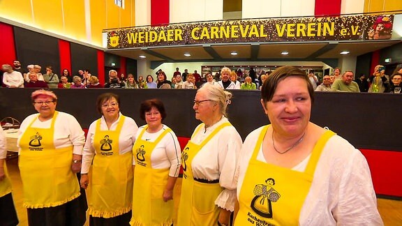 Eine Gruppe Frauen mit gelben Schürzen steht in einem Saal.