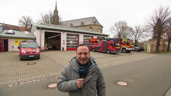 Ein Mann mit Mikrofon steht auf einem Platz mit Feuerwehrfahrzeugen.