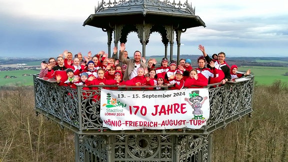 Ein Mann im Anzug und viele Menschen in rot-weißen Trainingsanzügen winken von einem eisernen Turm.