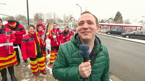 Ein lächelnder Mann mit Mikrofon steht vor einem Spielmannszug.