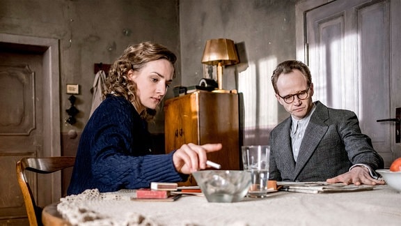 Erich (Christian Nähte) und Margot Honecker (Karolin Teska) sitzen an einem Tisch. Sie raucht eine Zigarette, er liest Zeitung.