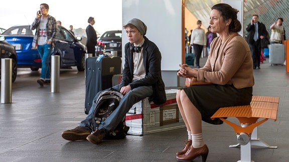 Eine Frau und ein Teenager sitzen vor dem Eingang eines Flughafens.