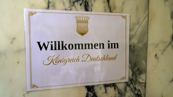 Ein Schild mit der Aufschrift „Willkommen im Königreich Deutschland“ hängt an einer Wand.