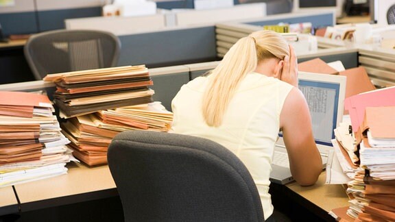 Eine Frau hält sich den Kopf an einem überfülltem Schreibtisch