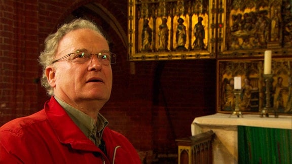 Ein Mann in roter Jacke steht vor einem reichverzierten Altar.