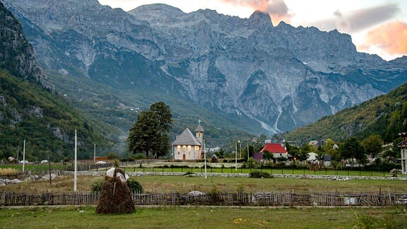 Panoramablick auf ein Gebirgsmassiv der albanischen Alpen, im Vordergrund eine kleine Kirche mit leuchtenden Fenstern