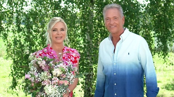 Eine Frau mit einem Blumenstrauß in der Hand und ein Mann stehen auf einer grünen Wiese vor einem Baum.