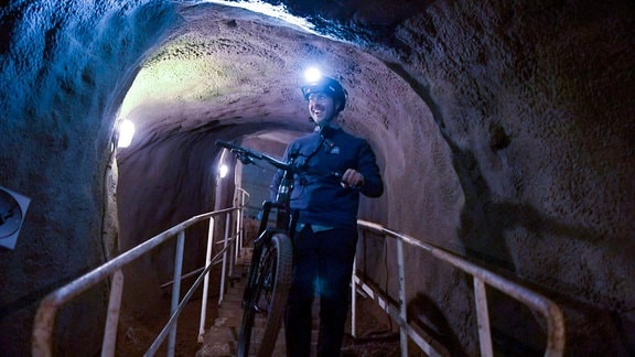 Ein Mann mit Stirnlampe und Fahrrad steht auf einer Brücke, die durch einen engen Tunnel führt.