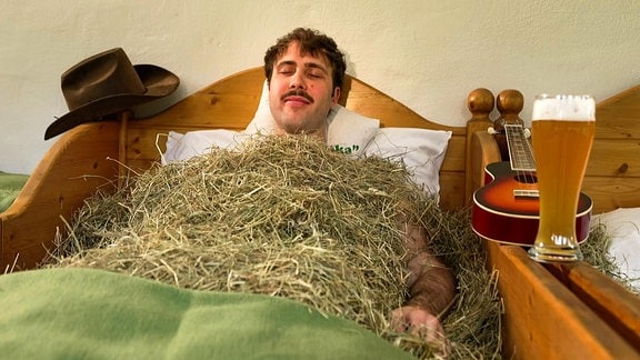 Ein Mann liegt mit Stroh bedeckt in einem Bett.