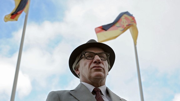 Blick von unten auf einen Mann mit grauem Anzug, Hornbrille und Hut mit mittelgroßer Krempe. Über ihm flattern Flaggen der DDR.