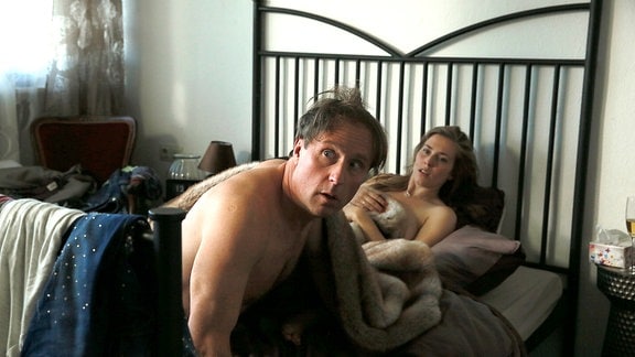 Ein unbekleidetes Paar sitzt in einem Bett.