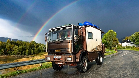 Ein kleiner LKW mit einer Wohneinheit steht auf einer Straße vor einem doppelten Regenbogen am Horizont.
