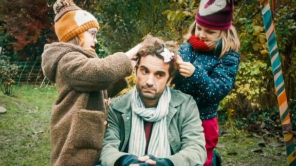 zwei Kinder flechten Schleifen in die Haare eines Mannes