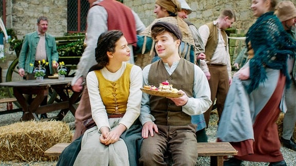 Mädchen und Junge sitzen auf einer Bank während eines Festes, Junge hält einen Teller in der Hand
