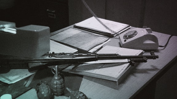 auf einem Schreibtisch liegen ein Gewehr und Handgranaten