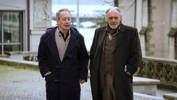 Zwei ältere Männer mit langen Wintermänteln auf einer Straße.