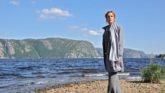 Eine Frau in offenem Mantel und losem Schal steht auf einem Strand gegenüber von hohen Felsen.