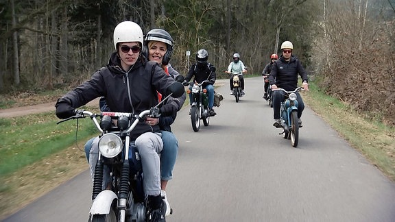 Eine Gruppe von Motorradfahrern kommt einen asphaltierten Weg durch einem Wald gefahren.