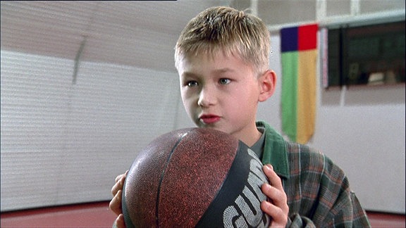 Ein Junge steht mit einem Basketball in einer Sporthalle.