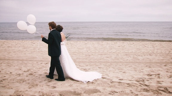 Ein junges Brautpaer spatziert am Strand mit weißen Luftballons