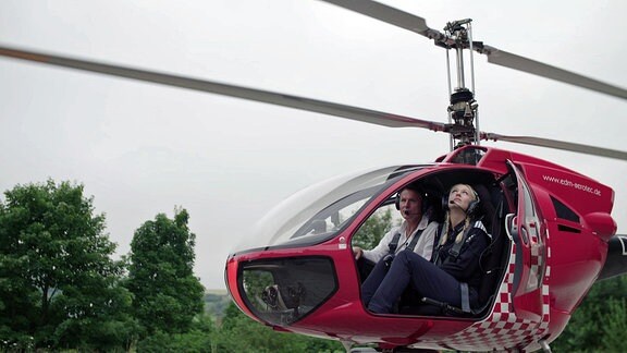 Ein Mann und eine Frau sitzen in einem kleinen, roten Hubschrauber. Die Frau schaut nach oben.
