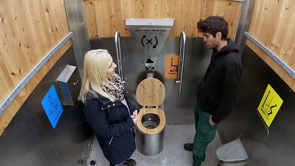 Eine Frau und ein Mann stehen in einem Toilettenraum und sprechen miteinander.