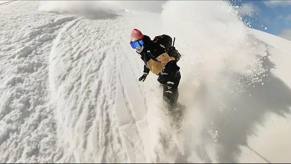 Ein Mann mit blauer Skibrille kommt auf einem Snowboard einen Berg hinunter und wirbelt Schnee auf.