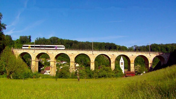 Eine Zug fährt über ein Viadukt, das über ein grünes Tal führt.