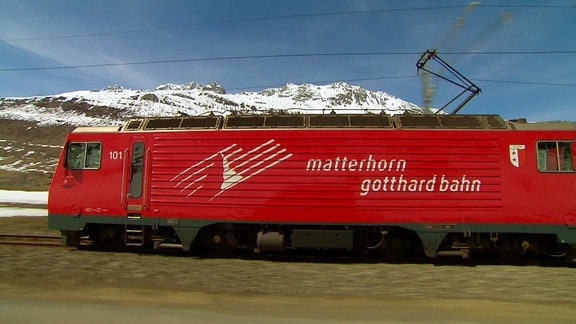 Eine rote Lokomotive fährt an einem schneebedeckten Gebirge vorbei.