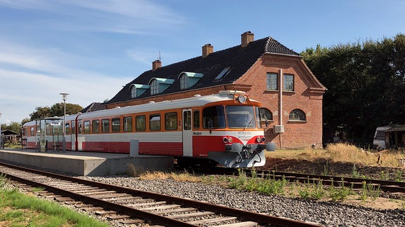 Ein rot-weißer Zug steht vor einem Backsteinhaus an einem Bahnsteig.