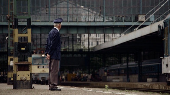 Ein Mann in Eisenbahneruniform steht an einem Bahnsteig in einem alten Bahnhof.
